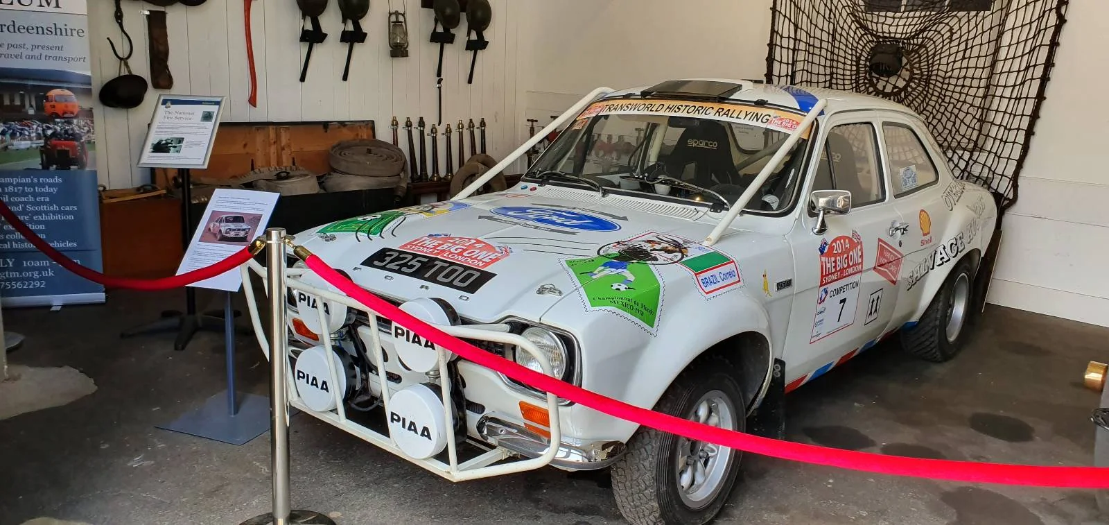 A vintage white rally car with various sponsor stickers is displayed in a museum setting. It's behind red velvet ropes, with informational panels and decorative items on the wall, including old tools and a net.