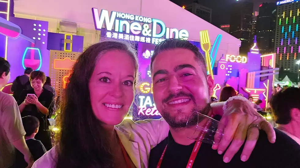 Smiling pair at Hong Kong Wine & Dine Festival. Neon backdrop with "FOOD" sign. Crowded setting, vibrant colors, festive mood.