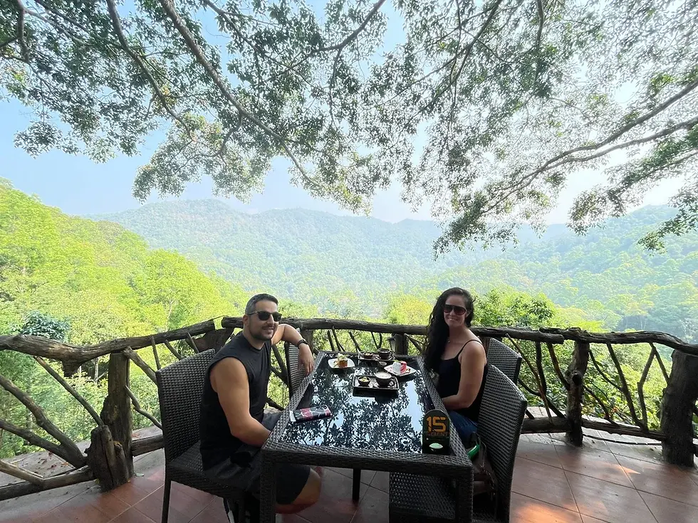 Two people sitting at an outdoor table with food, overlooking lush green mountains. Both are wearing sunglasses, creating a relaxed vibe.