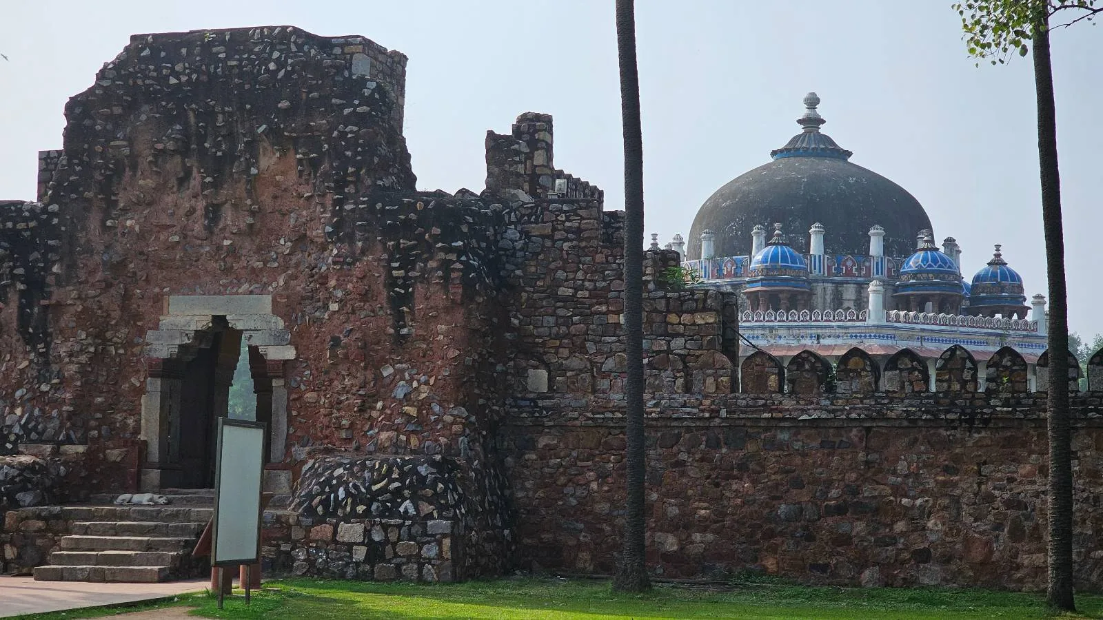 Ancient stone fortress wall with an arched entrance on the left. Behind it, a large dome with smaller blue and white domes is partially visible. Two tall palm trees frame the scene, set against a clear sky.
