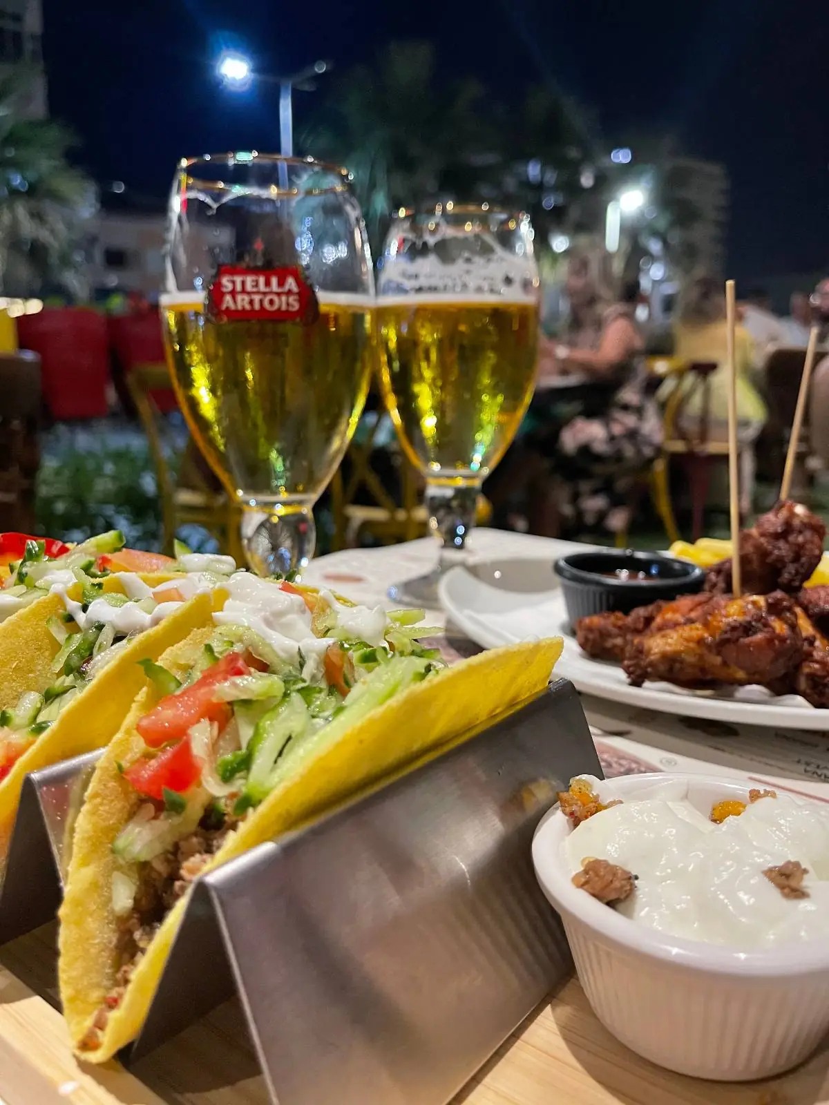 Three tacos filled with vegetables and meat are served on a metal stand, accompanied by a bowl of sauce, chicken wings, and two glasses of beer on an outdoor table at night.