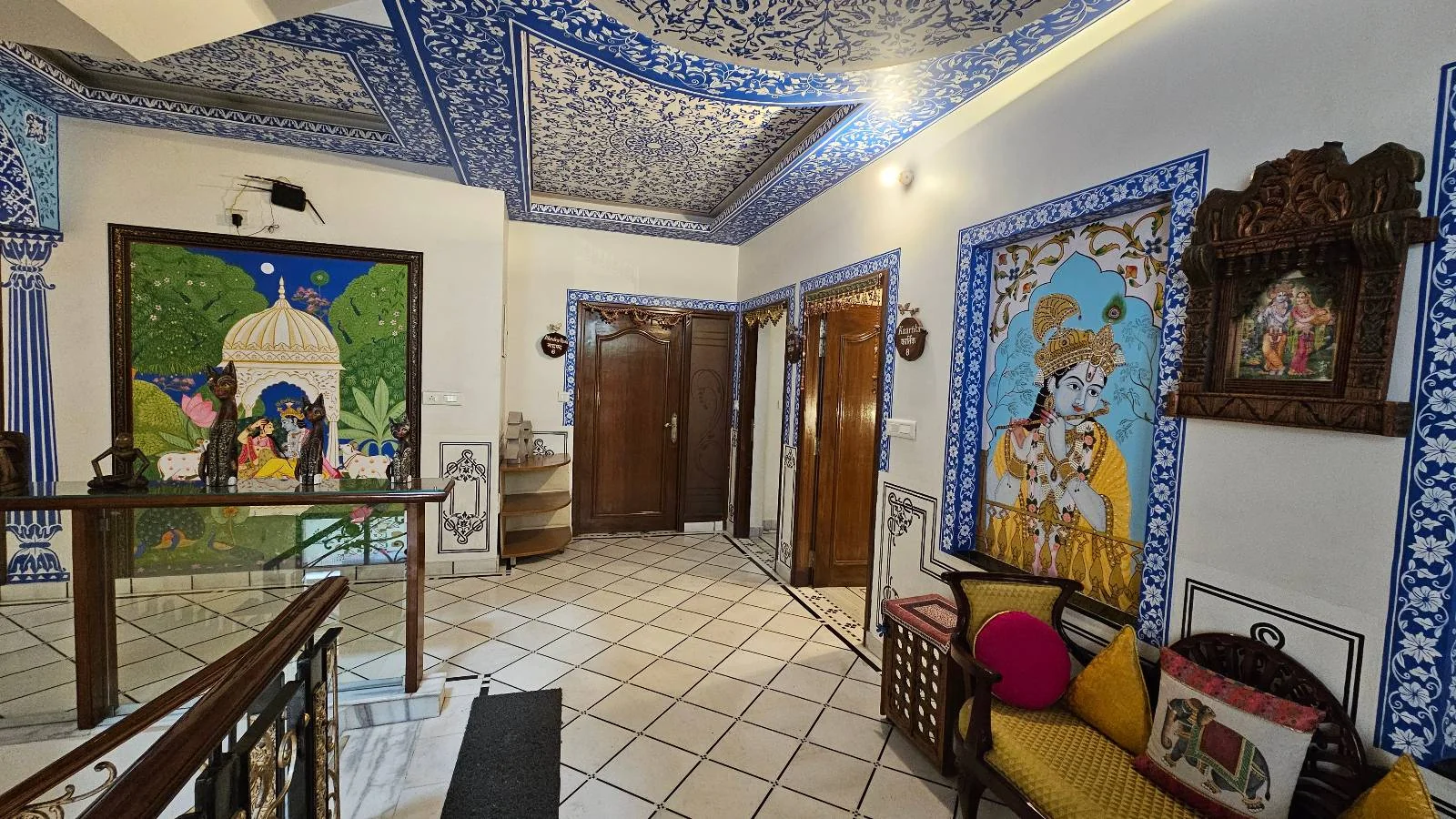 A vibrant hallway features traditional Indian artwork, blue patterned ceiling, tiled floor, wooden doors, a cushioned bench with colorful pillows, and decorative wall portraits in ornate frames.