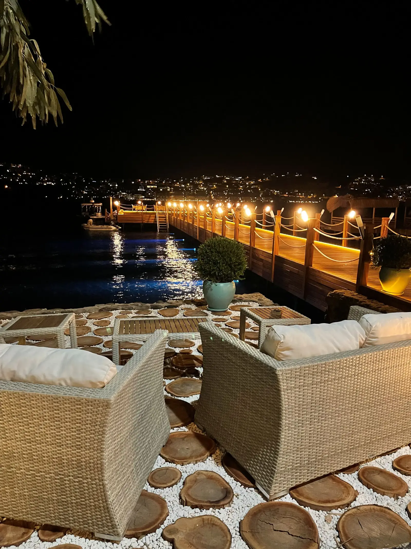 Two wicker chairs on a patio overlook a lit pier extending into the water at night, with distant city lights in the background.