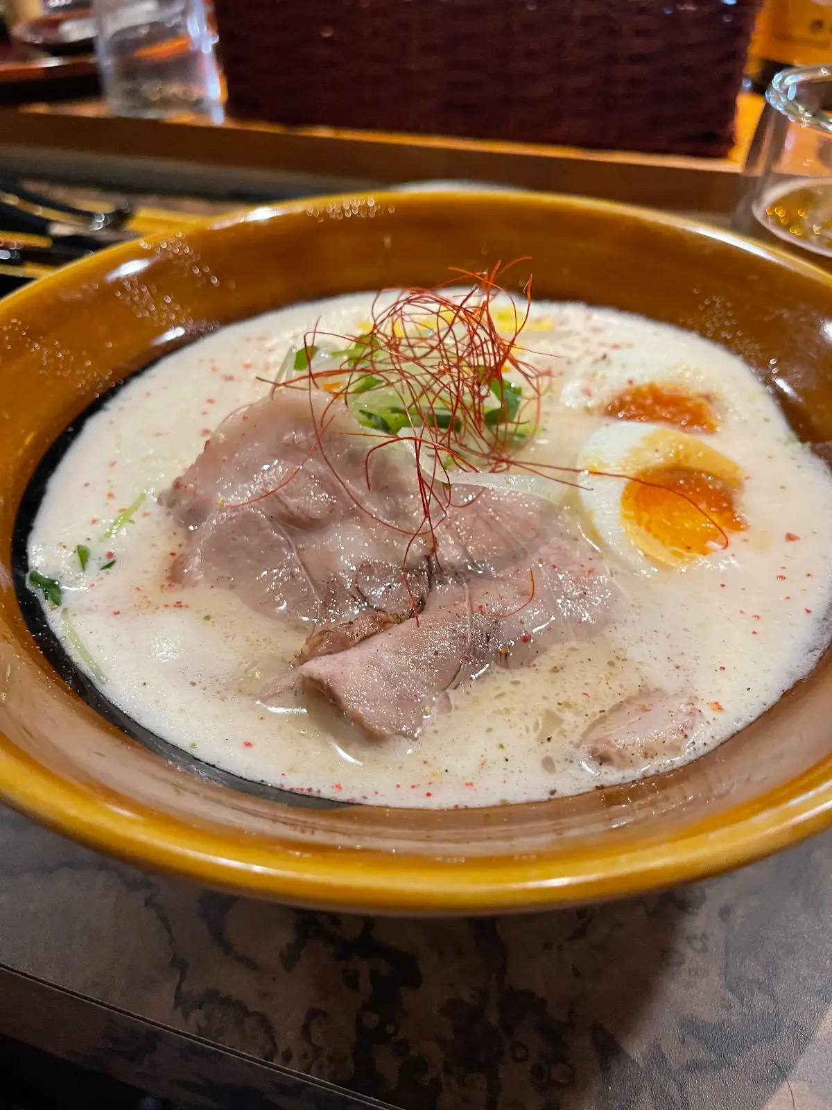 A bowl of ramen with creamy broth, slices of pork, half a soft-boiled egg, and garnished with green onions and thin red chili threads.