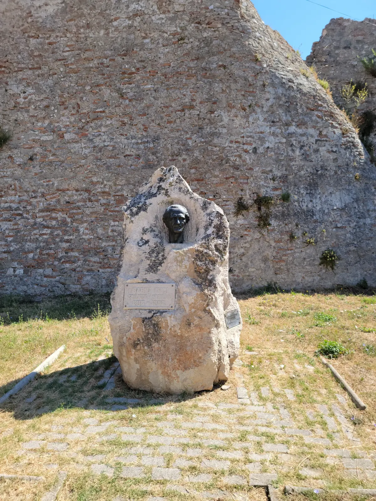 A bronze bust is mounted on a large stone with a plaque, set outdoors on a paved area in front of an old, weathered stone wall. Grass and blue sky are visible in the background.
