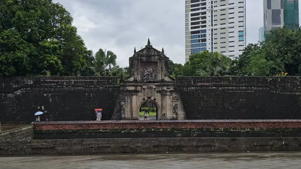 Historic stone gate with greenery, people holding umbrellas. Tall modern buildings in the background, overcast sky. Calm, historic ambiance.