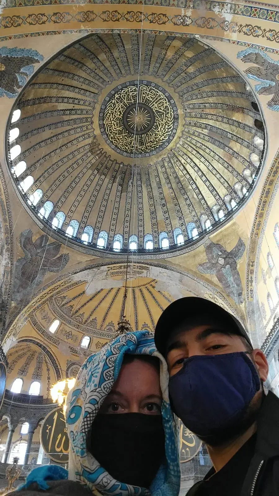 Two masked people in a mosque with a large ornate dome above. The dome features intricate patterns and Arabic calligraphy.