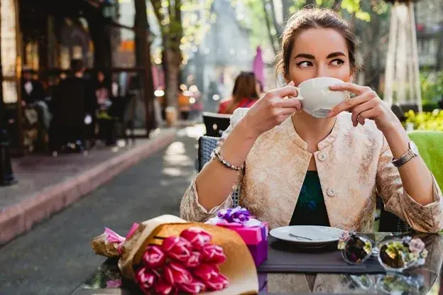 Woman sipping coffee at an outdoor café, surrounded by a gift and pink flowers. Sunlit street in the background, creating a relaxed vibe.
