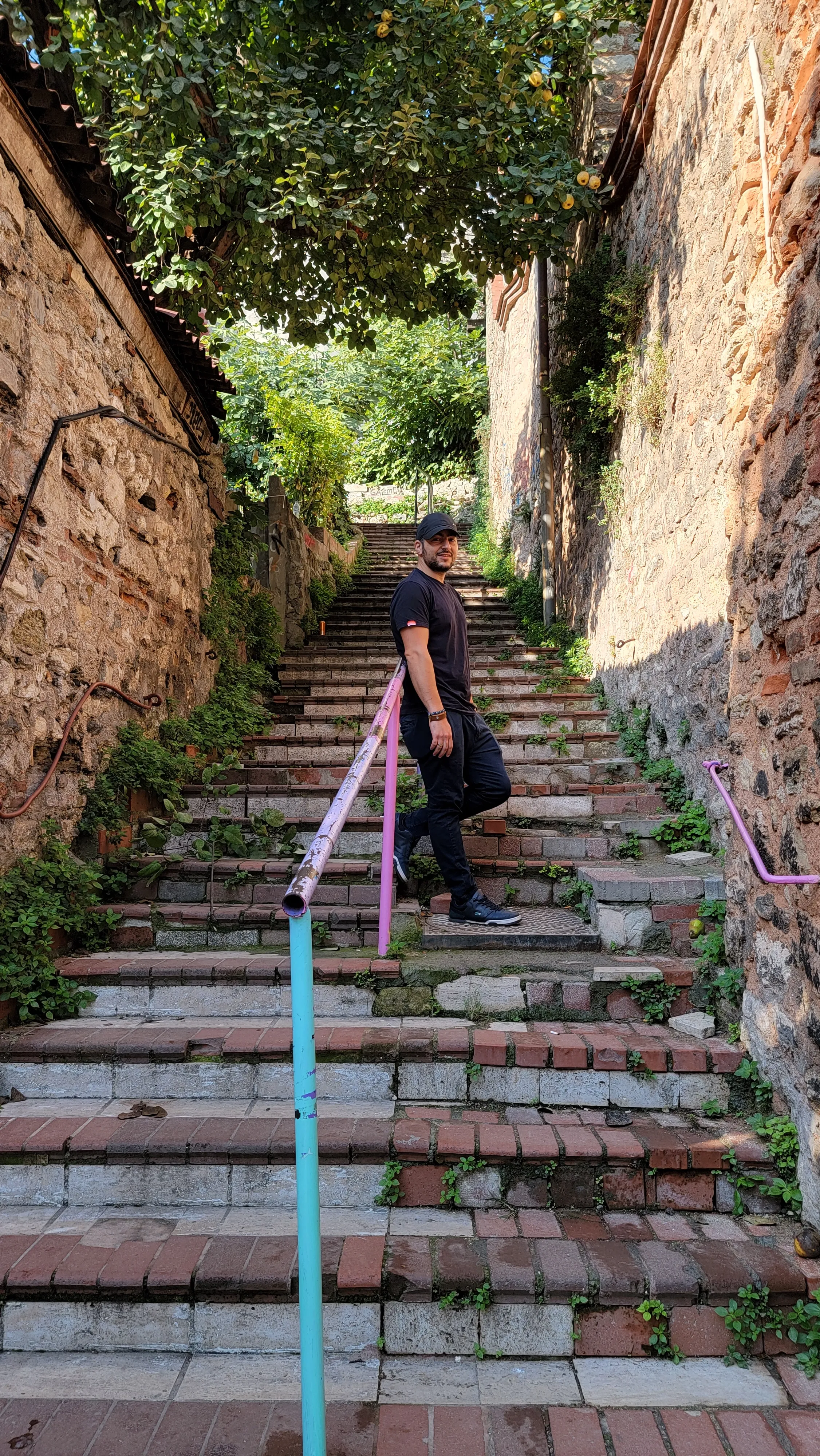 A person dressed in black walks up a narrow outdoor staircase bordered by old brick walls and greenery, holding a dark bag and looking back at the camera.