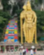 Golden statue beside vibrant steps at Batu Caves. People and pigeons in foreground, lush greenery and rocky cliffs in background.