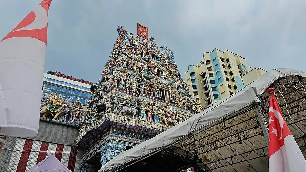 Colorful Hindu temple adorned with intricate statues, flanked by Singapore flags. Tall modern buildings in the background under a cloudy sky.