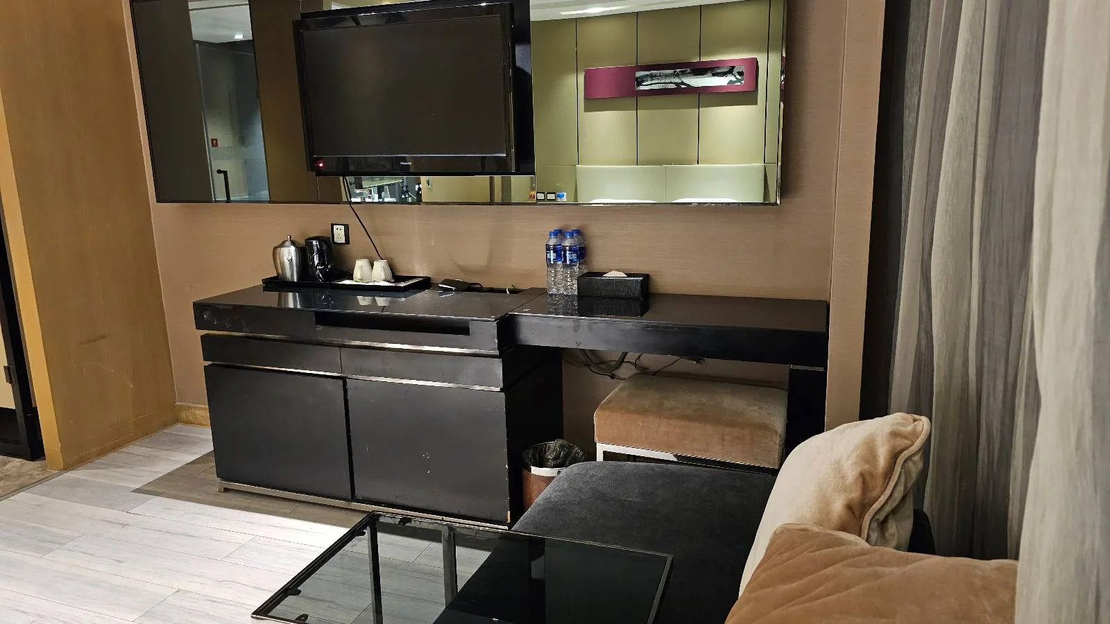 Hotel room corner with a black desk, a TV mounted above, a coffee and tea station, a mirror, a glass-top table, a beige cushion, and partially visible curtains on the right. Modern and tidy setup.