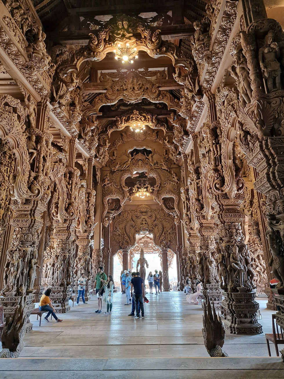 People explore a intricately carved wooden temple hall in bright light. The ornate arches and pillars feature detailed patterns and sculptures.
