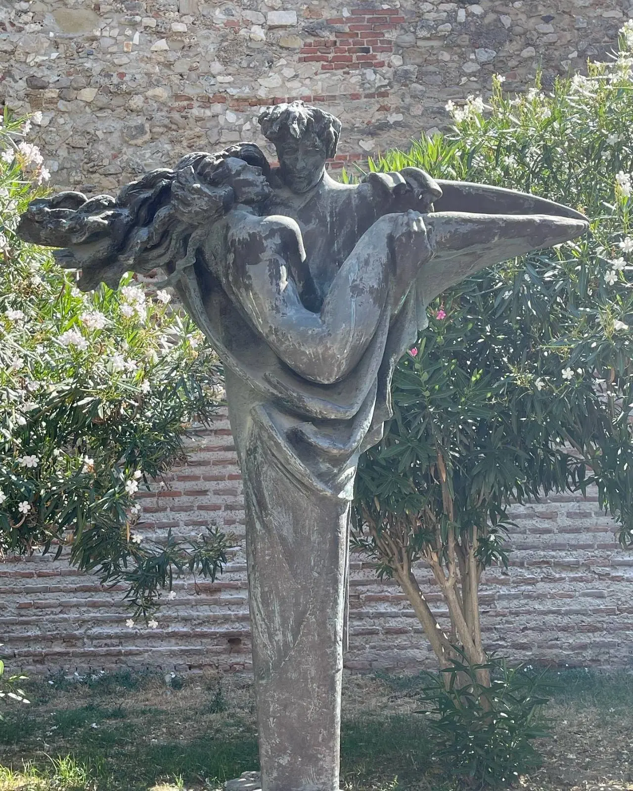 A bronze statue depicts a standing figure holding another figure horizontally in their arms, set outdoors with greenery and a brick wall in the background.