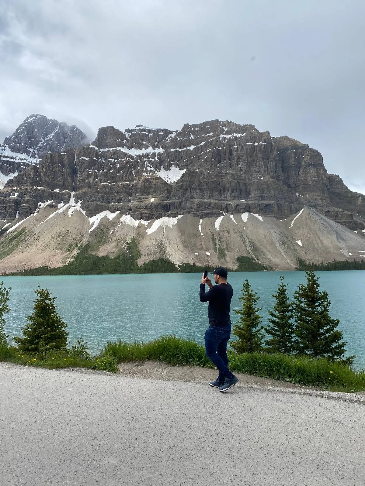 A person stands near a lakeshore taking a photo, with pine trees in the foreground and a large rocky mountain with patches of snow rising behind the turquoise water under a cloudy sky.
