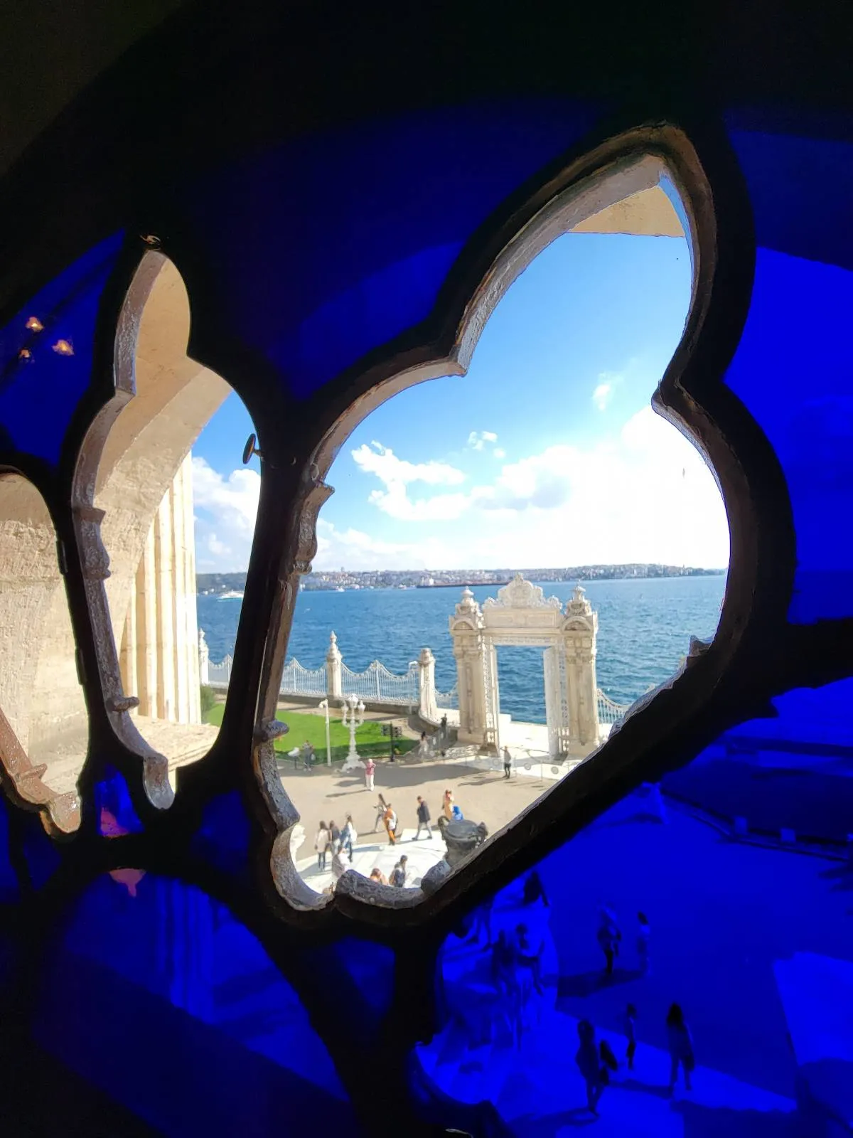 A view of a waterfront with blue sky and clouds, seen through a decorative, quatrefoil-shaped window with blue stained glass. Stone arches and greenery are visible outside by the water.