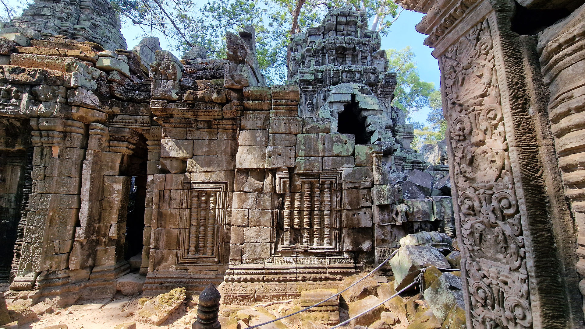 Ancient stone temple ruins covered in intricate carvings and surrounded by trees, with sunlight filtering through the foliage above. Some walls and structures are partially collapsed or weathered.