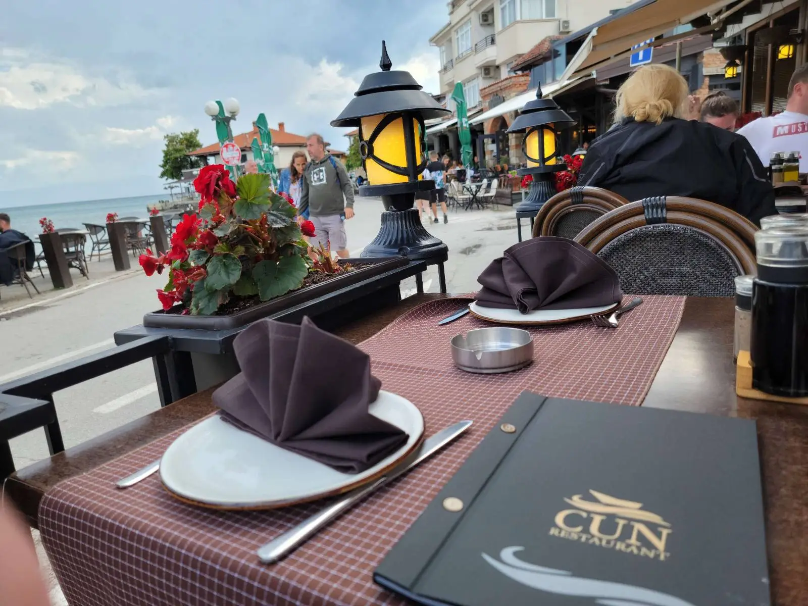 Outdoor restaurant scene with two table settings topped with dark napkins and a menu on a checkered tablecloth. A decorative lamp and a pot of red flowers are on the table. People and buildings are visible in the background under a cloudy sky.