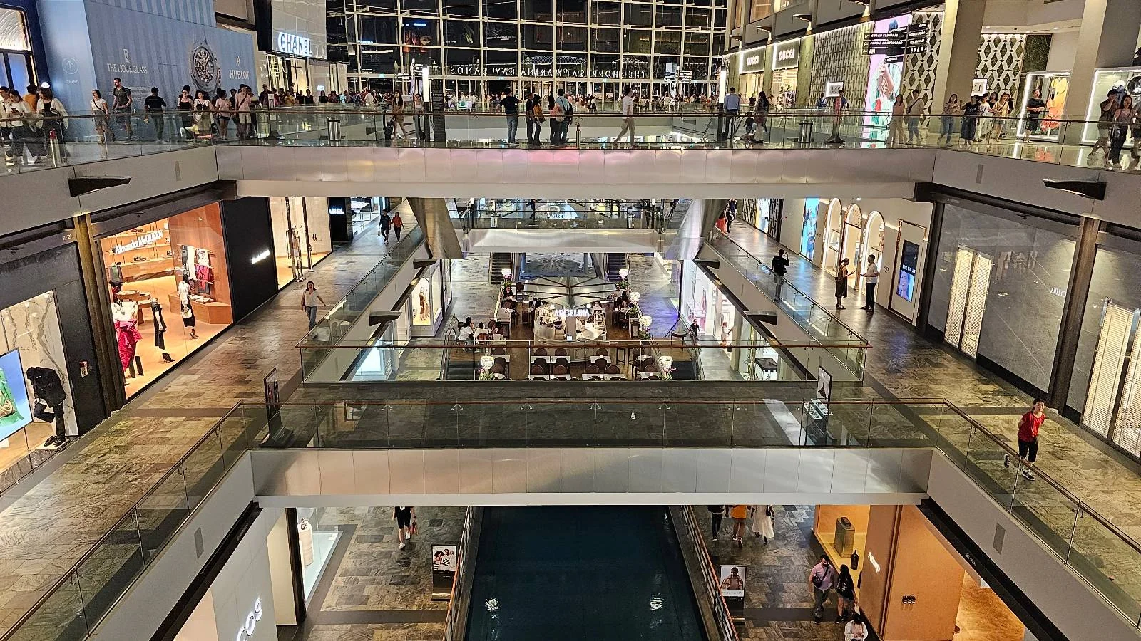 A multi-level shopping mall with glass railings and various storefronts. People are walking and shopping on both levels. The architecture features open spaces and modern design elements. Brightly lit with a relaxed atmosphere.