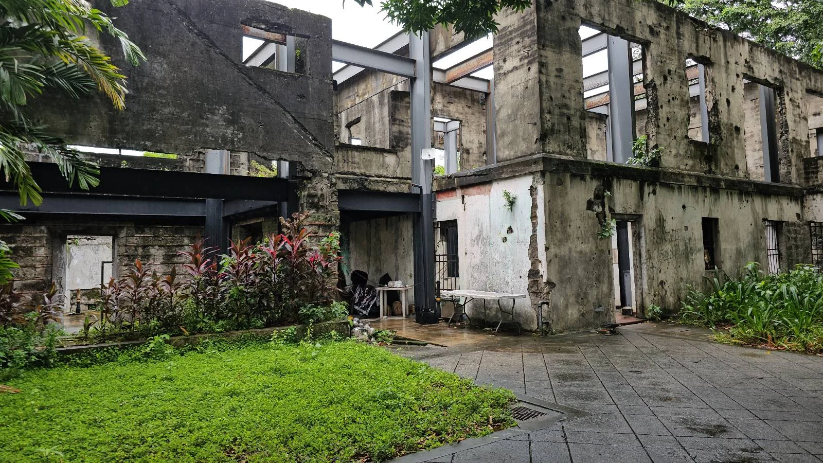 A weathered, partially roofless stone building surrounded by greenery. A small garden with pink flowers is in the foreground, and a pathway leads to the structure. The scene has an overcast ambiance.