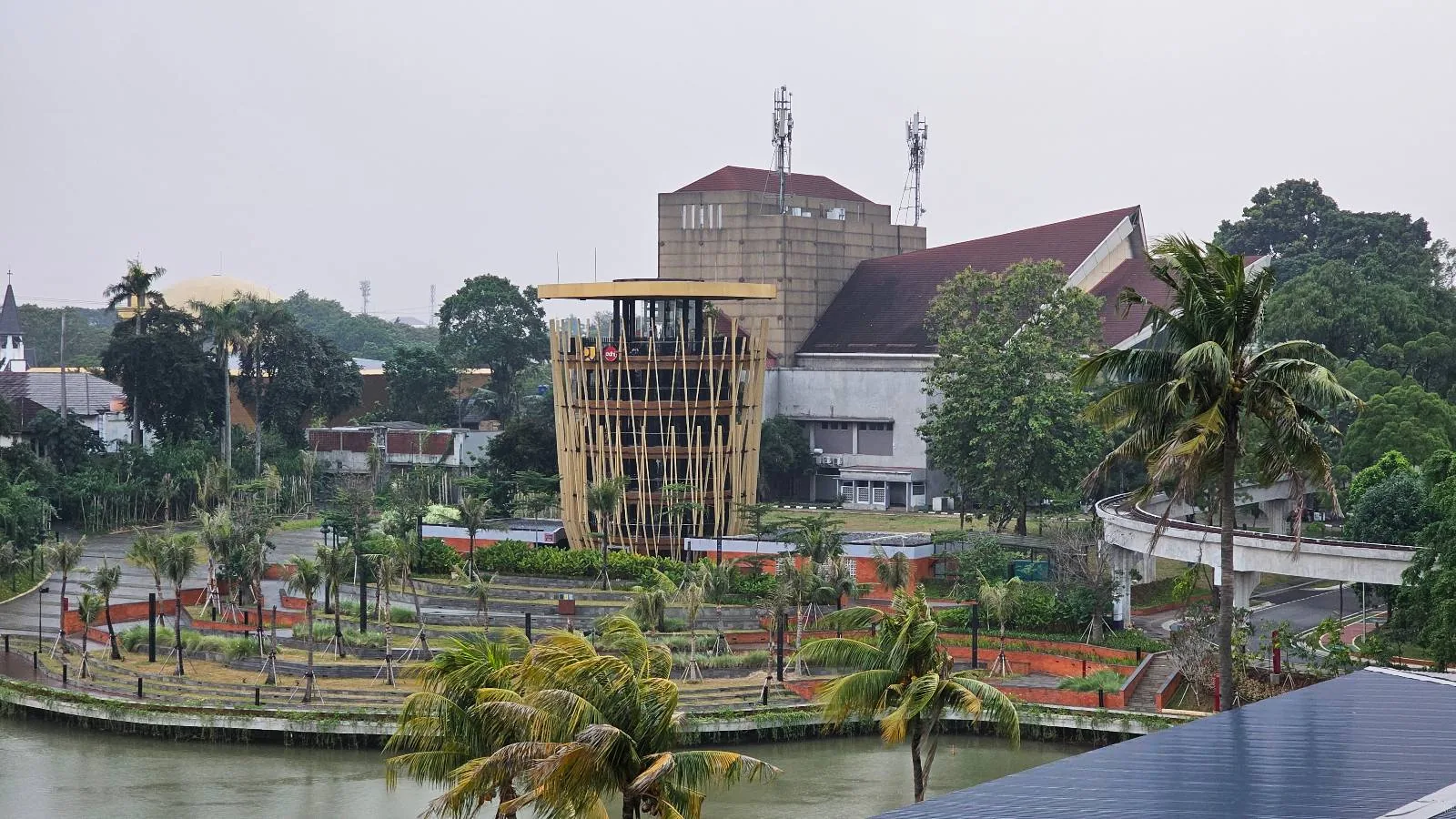 A circular bamboo structure stands in the center of a landscaped area surrounded by a water canal. Palm trees and manicured gardens are visible, with a large building and more greenery in the background under an overcast sky.