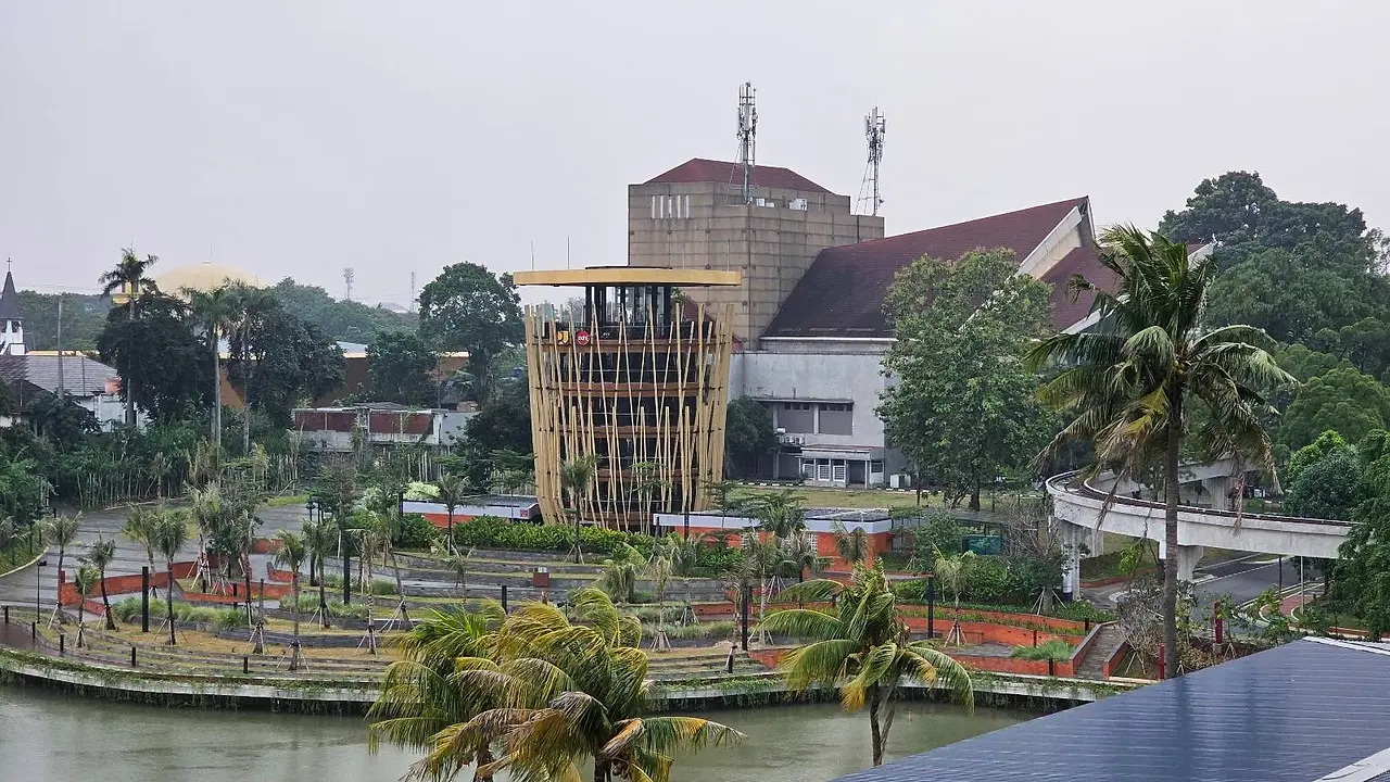 A circular bamboo structure stands in the center of a landscaped area surrounded by a water canal. Palm trees and manicured gardens are visible, with a large building and more greenery in the background under an overcast sky.