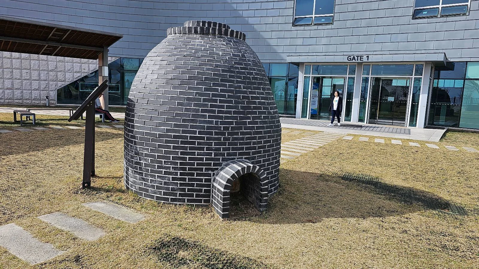 A brick kiln-shaped sculpture stands on a grassy area in front of a modern building with glass doors. A small arched opening is visible at the base of the structure. A person is entering the building in the background.