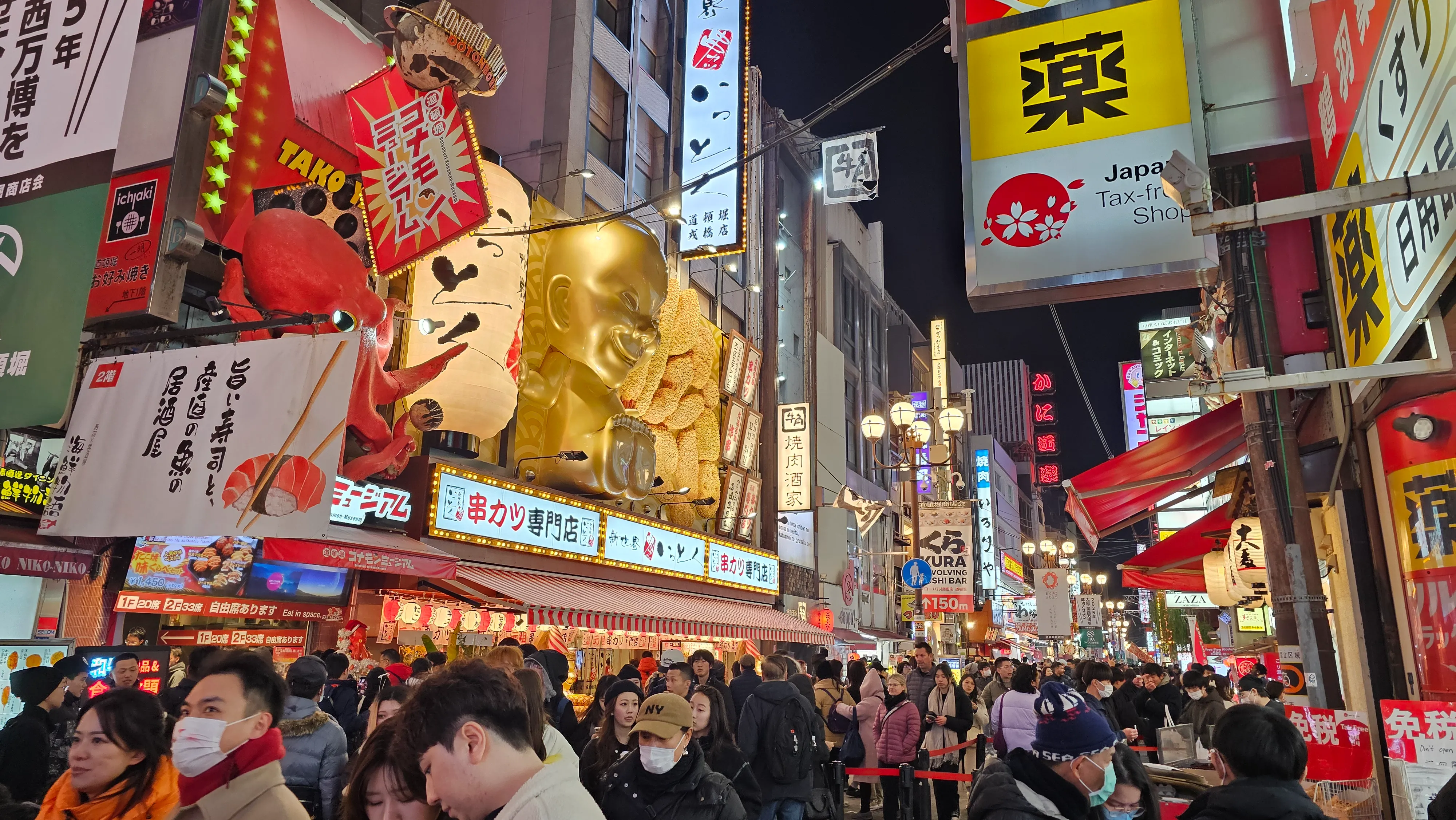 A busy street scene in Japan at night, filled with people, colorful signs, restaurants, and a prominent golden statue of a chef above a building. Neon lights and shop banners create a lively, bustling atmosphere.