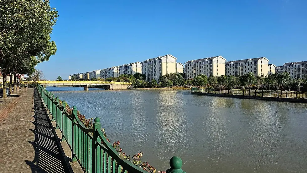 River with a green railing walkway on the left, yellow bridge, and apartment buildings in the background. Clear blue sky, calm scene.