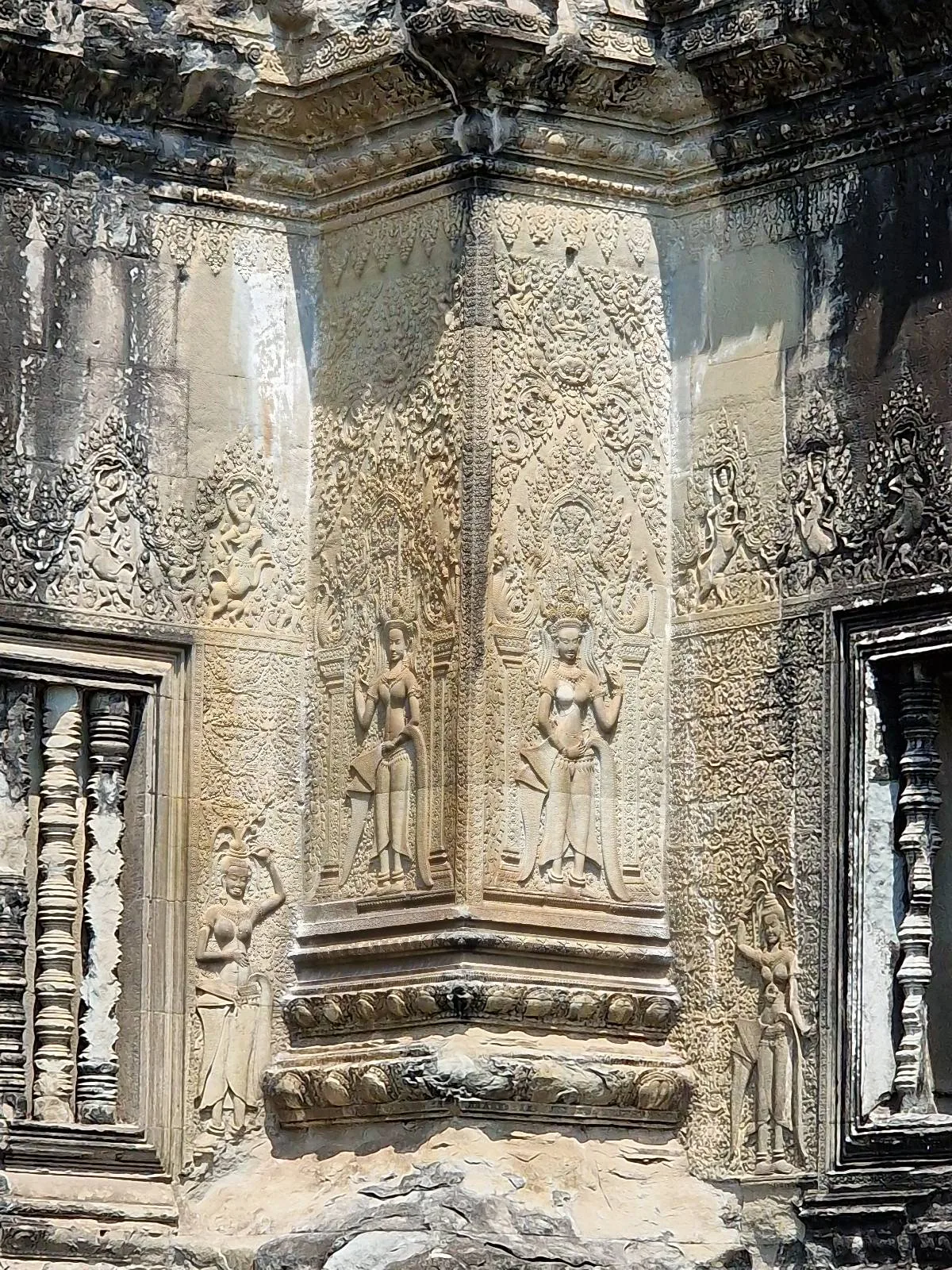 Detailed stone carvings of standing figures and ornate patterns decorate a section of an ancient temple wall, with small windows featuring stone balusters on either side.