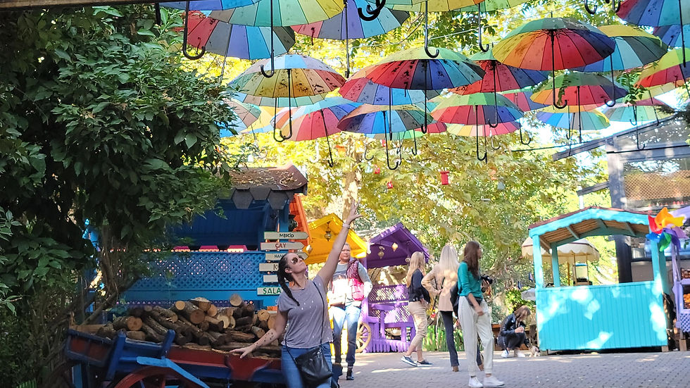 Woman reaching towards colorful umbrellas hanging in a park. Bright stalls and leafy trees in the background. Playful, joyful atmosphere.
