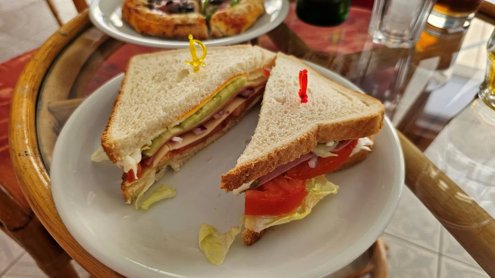A plate with a sandwich cut in half, filled with lettuce, tomato, onions, and deli meat, secured with colored toothpicks. A blurred plate of food, glass, and drink are in the background.