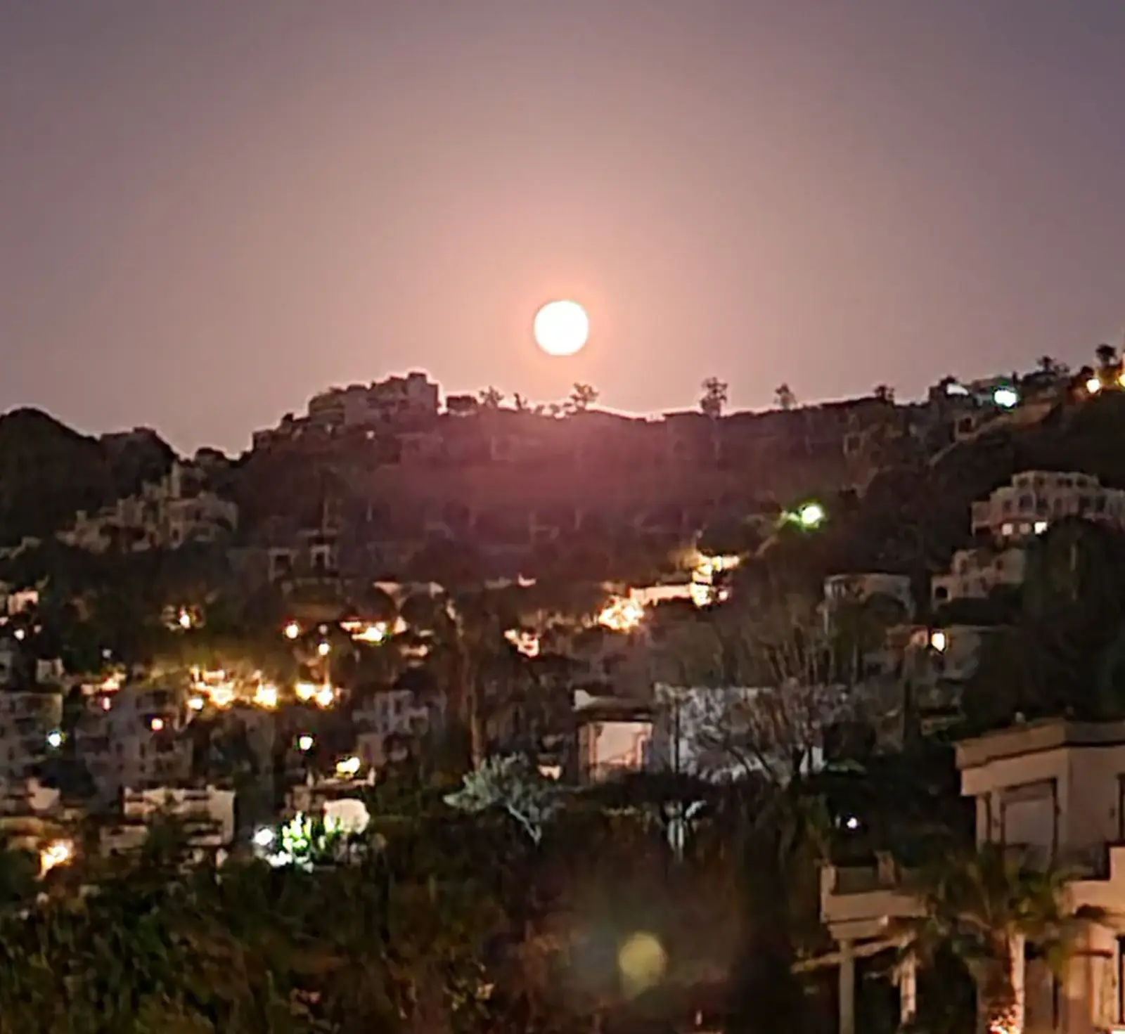 Full moon rising over a hillside town with scattered lights illuminating buildings and trees.