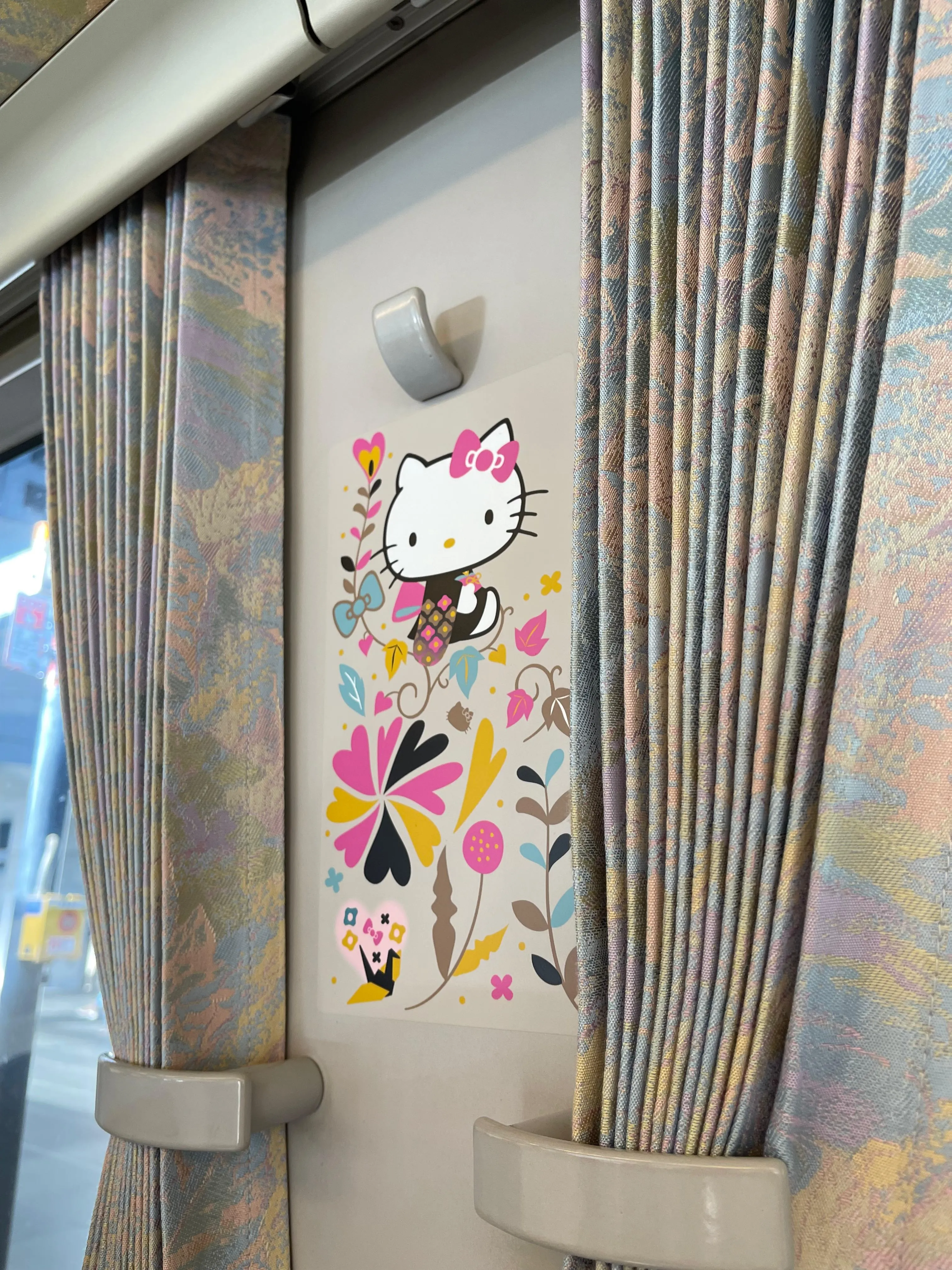 A small Hello Kitty decoration with flowers and leaves hangs on a wall between patterned curtains inside a vehicle, possibly a bus or train.