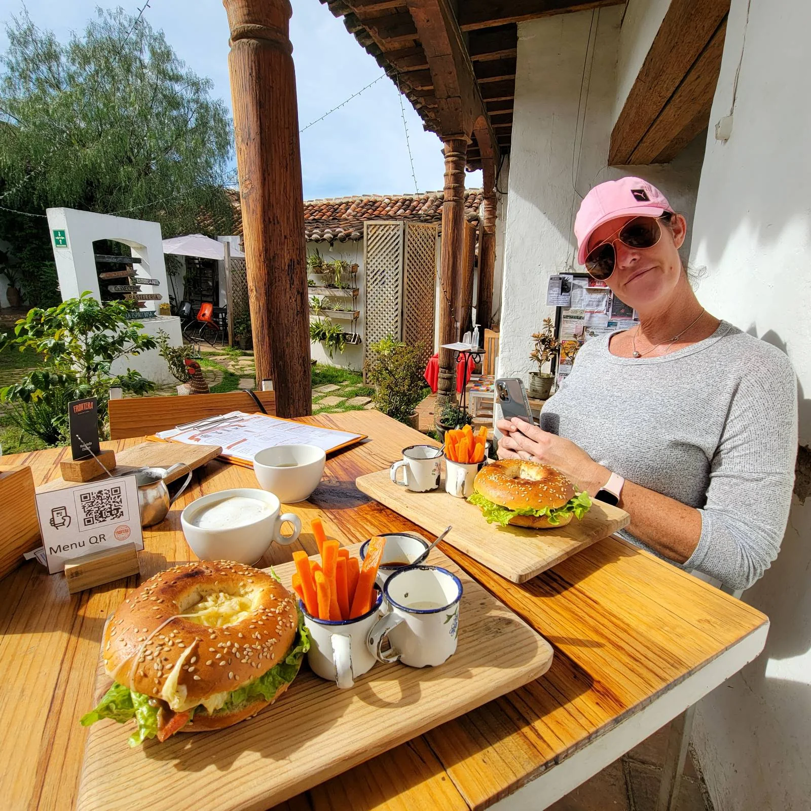 A person wearing sunglasses and a pink cap sits at an outdoor table with two bagel sandwiches, carrot sticks, and dips on wooden trays. The setting is sunny with greenery and wooden beams in the background.