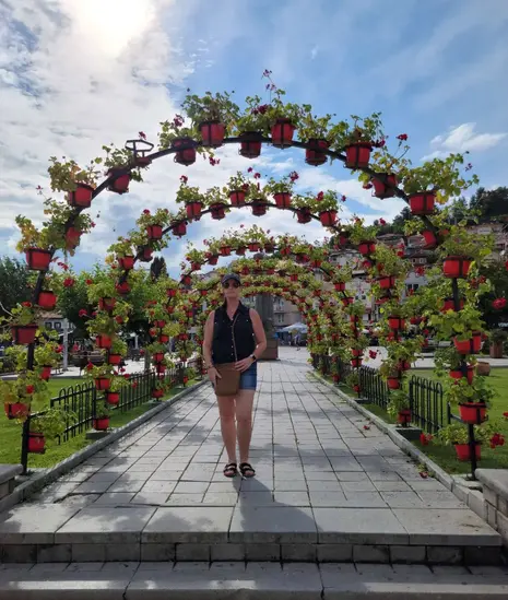 A person stands on a stone walkway beneath a potted archway adorned with red flower pots and lush greenery. The setting is a park on a sunny day, with picturesque clouds in the sky and a hillside in the background, perfect for photo ops.