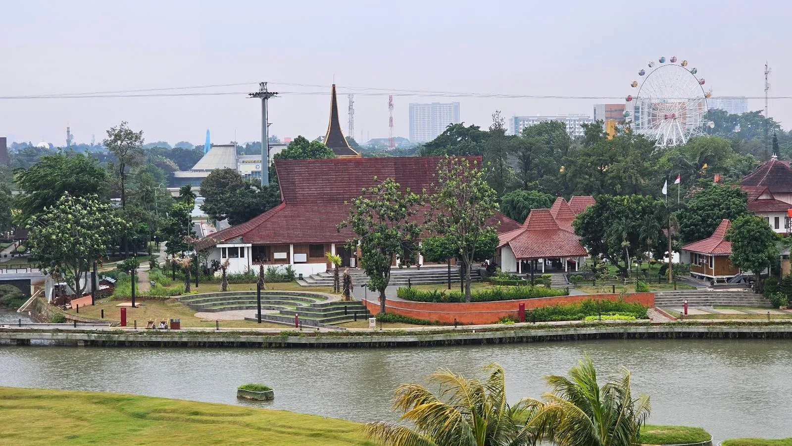 A scenic view of Taman Mini Indonesia Indah in Jakarta, featuring traditional Indonesian buildings with brown roofs, surrounded by lush greenery and a river in the foreground. A Ferris wheel and cityscape are visible in the background.