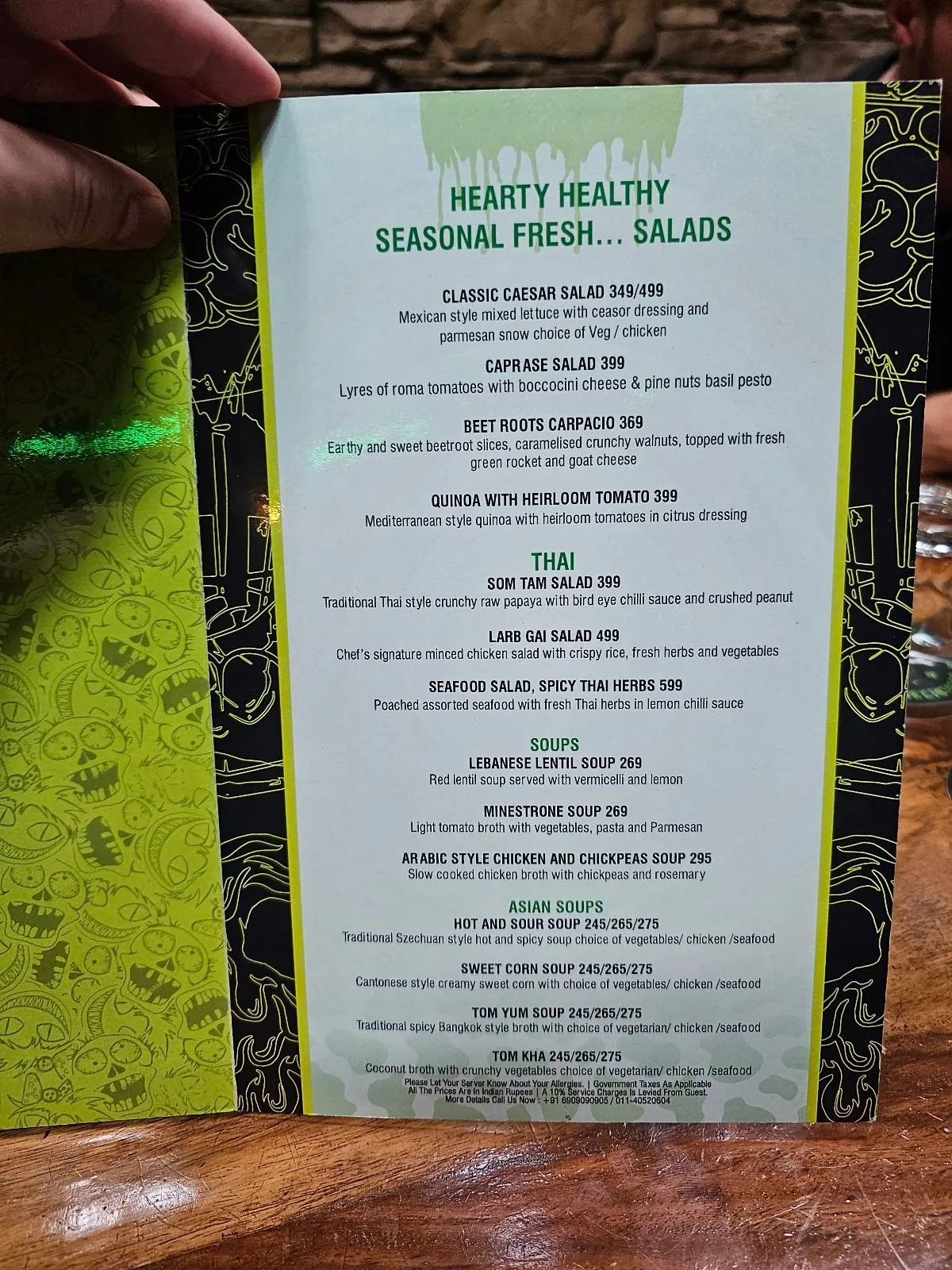 A close-up of a restaurant menu titled "Hearty Healthy Soothing Fresh Salad," listing various salad options in white text on a green and black decorative background, with a hand holding the menu open.