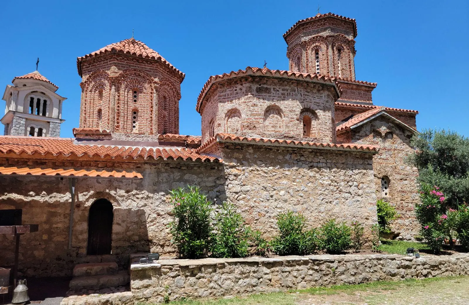 Ancient stone church with red-tiled roofs and intricate brickwork, set against a bright blue sky. Green shrubs and a small garden surround the building.