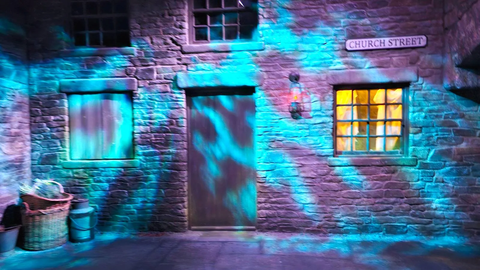 A brick building with a closed door, a window with yellow light, a basket and bin outside, and turquoise-blue light patterns projected onto the wall at night.