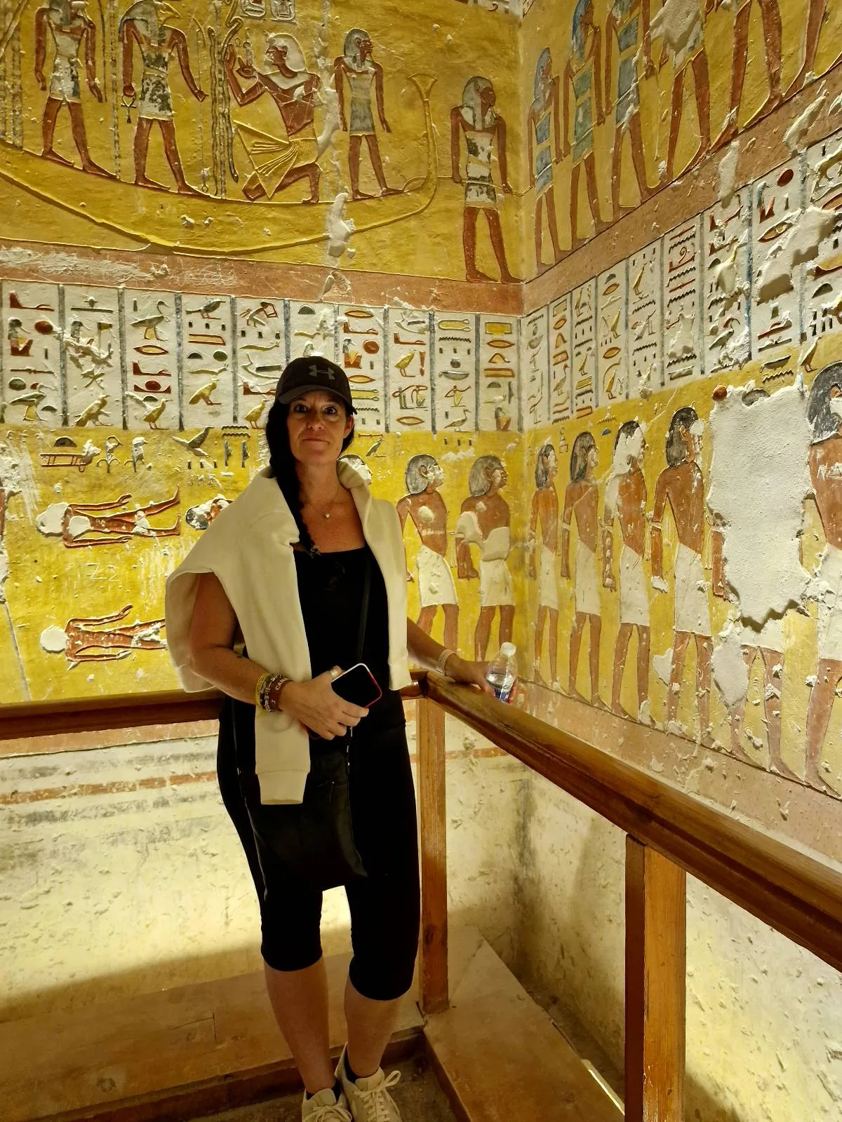 A person stands inside an ancient Egyptian tomb, surrounded by vibrant hieroglyphics and wall paintings depicting various figures. The person holds a phone and wears casual black and white clothing.