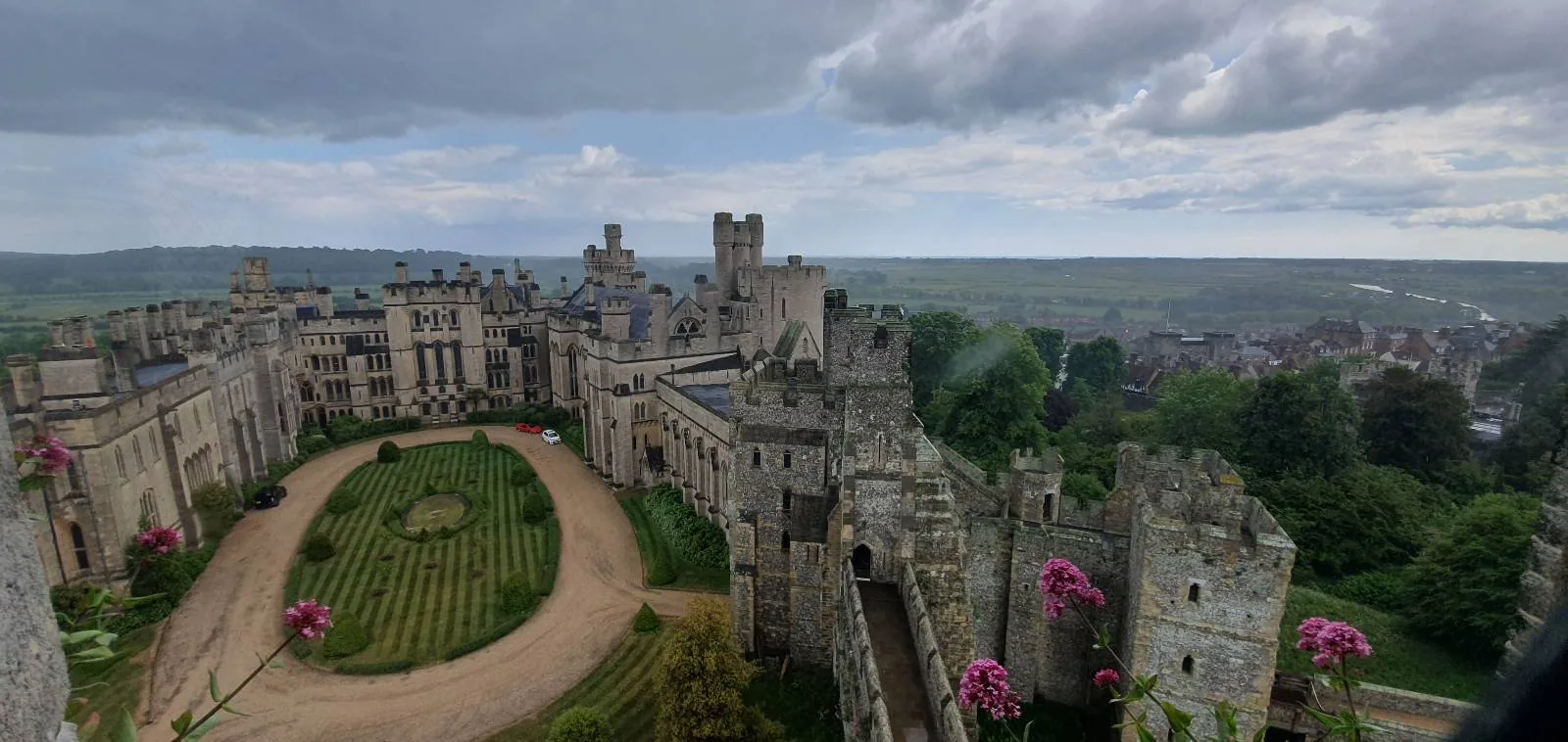 A vast, historical castle with gray stone walls and numerous turrets under a partly cloudy sky. A neatly landscaped garden with a circular path and trimmed hedges is in the foreground, surrounded by lush greenery and rolling hills.