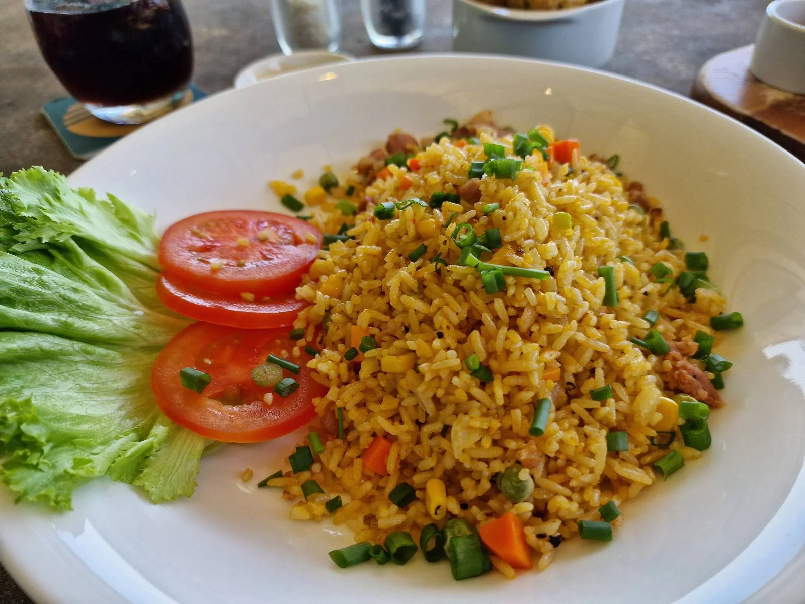 A plate of fried rice garnished with chopped vegetables and herbs, accompanied by two slices of tomato and a few lettuce leaves. In the background, there is a glass of dark beverage and small bowls of condiments.