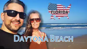 A smiling pair in sunglasses at Daytona Beach with a USA map featuring "Florida" in stars and stripes. Waves and blue sky in background.
