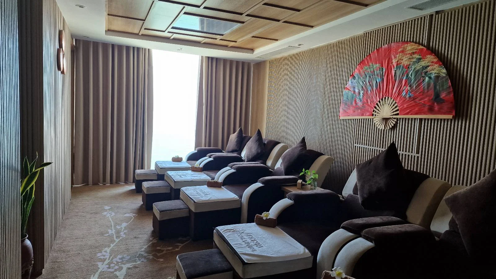A serene spa room features multiple plush recliners with footrests, arranged in a row. Soft lighting and a large decorative fan on the back wall create a calm ambiance. Draped curtains partially cover a large window, enhancing the peaceful atmosphere.
