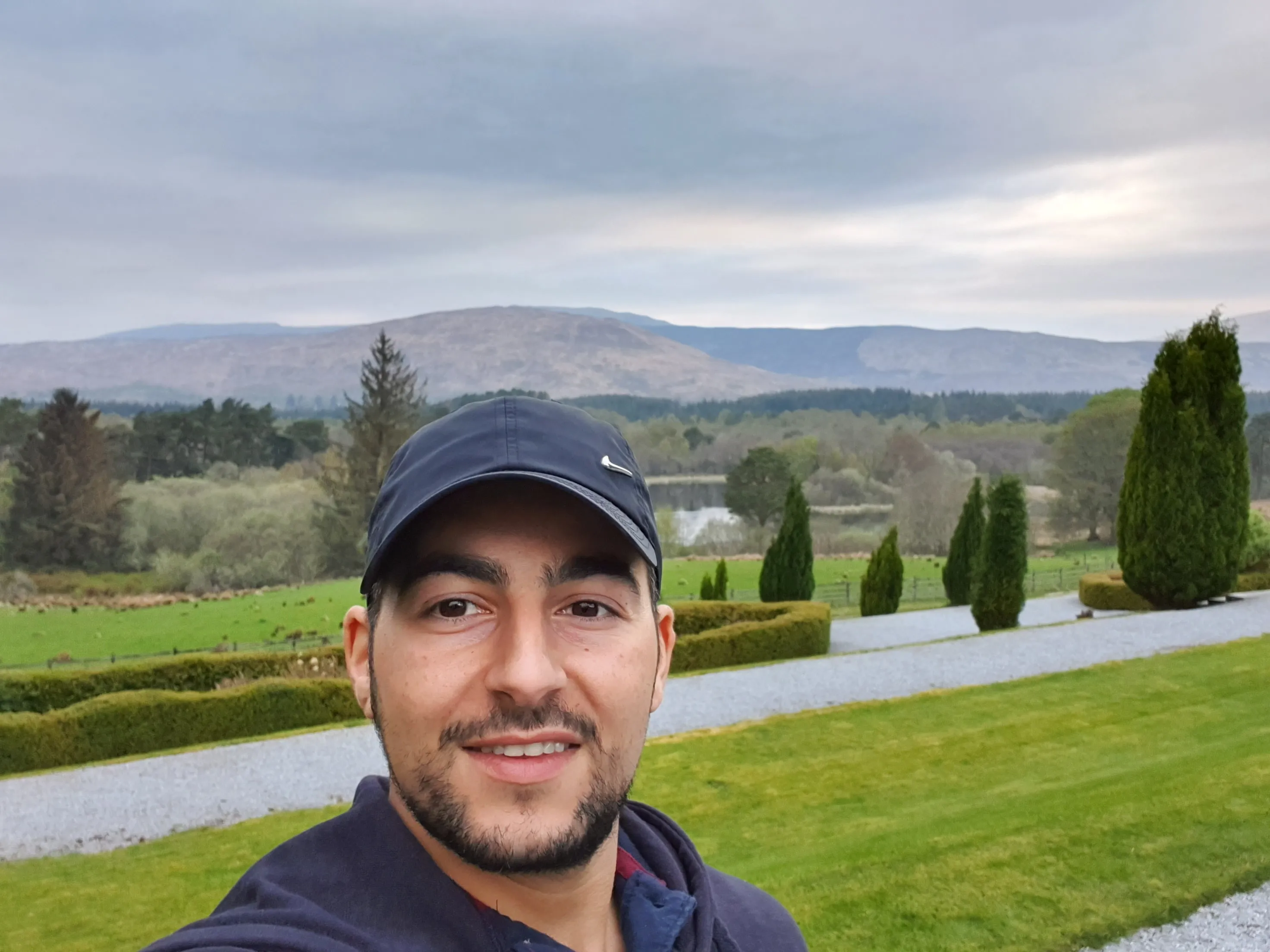 A man in a dark baseball cap smiles for a selfie with the green lawn and distant hills of the Scottish Highlands behind him.