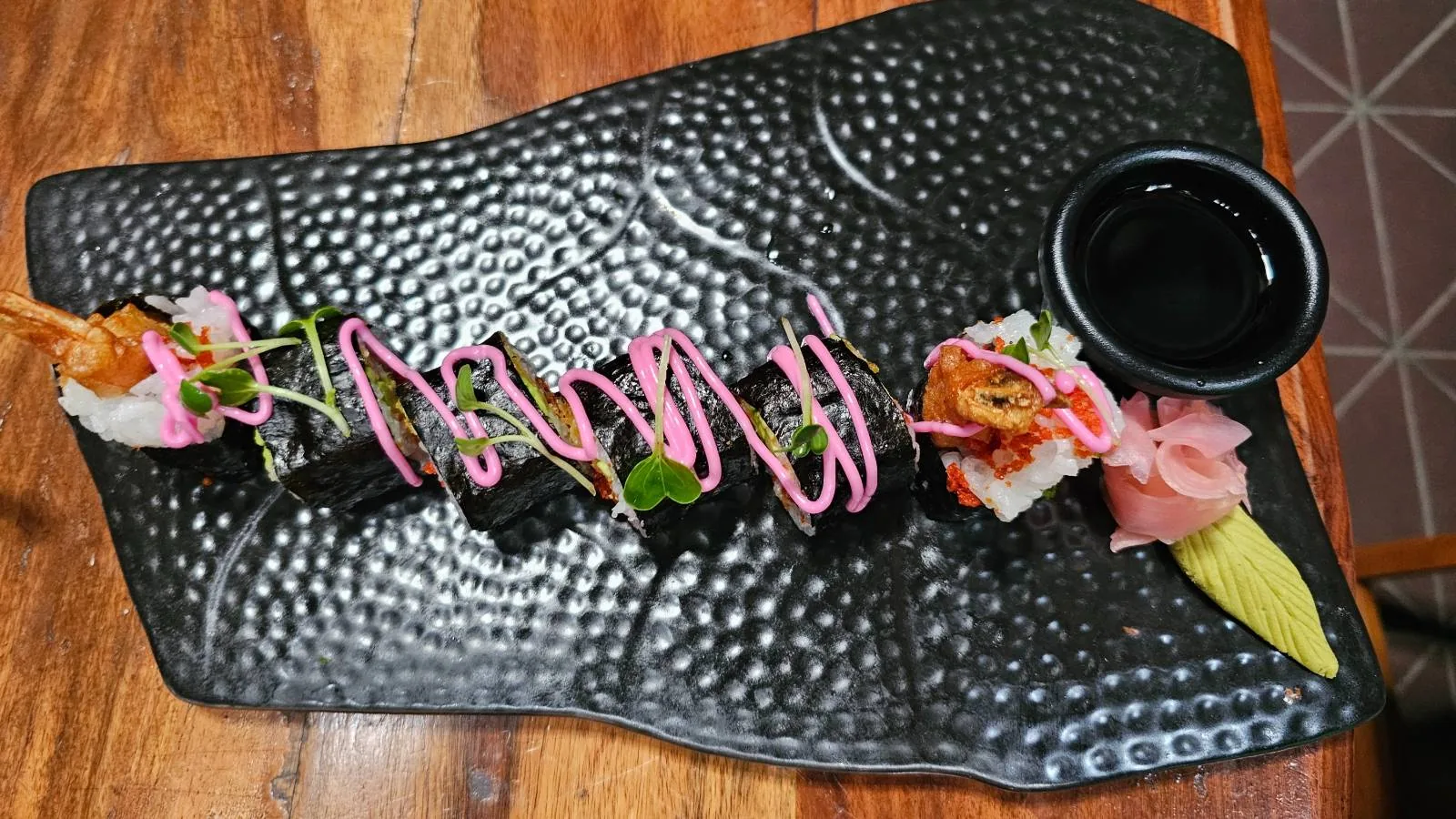A rectangular black plate holds sushi rolls garnished with pink sauce and shrimp, accompanied by pickled ginger, wasabi, and a small dish of soy sauce. The plate is on a wooden table.