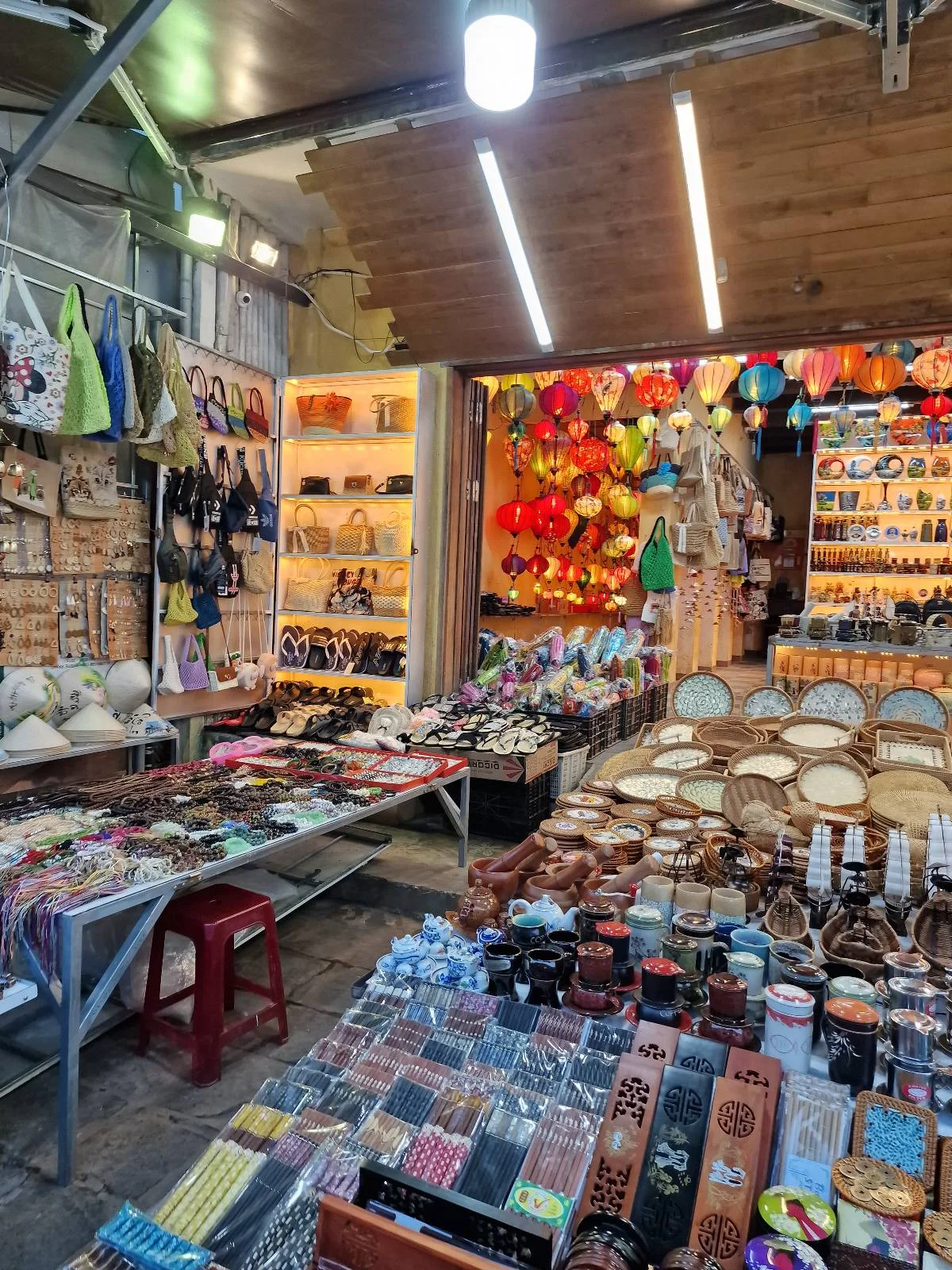 A vibrant market stall displays colorful bags, hats, jewelry, fans, and souvenirs neatly arranged on tables and shelves under bright lighting.