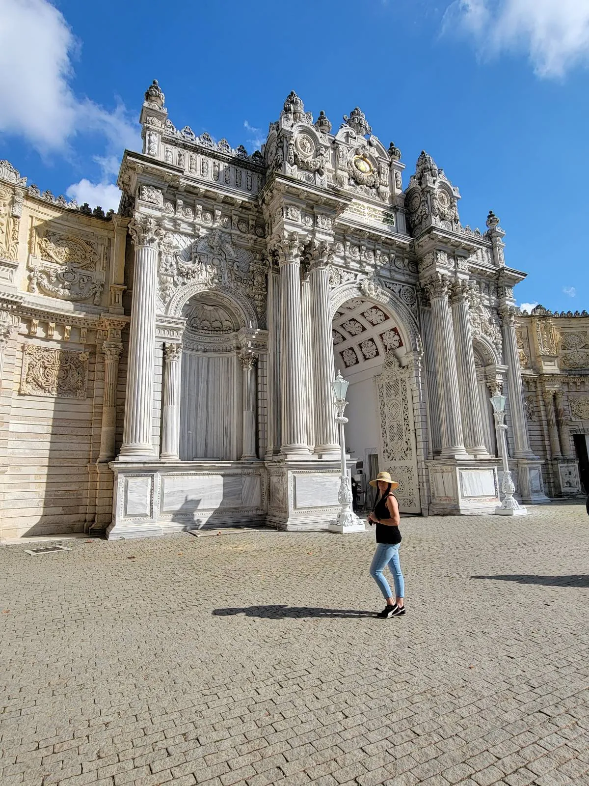 Person walking in front of an ornate, historic stone gate with intricate carvings and columns, set against a clear blue sky.