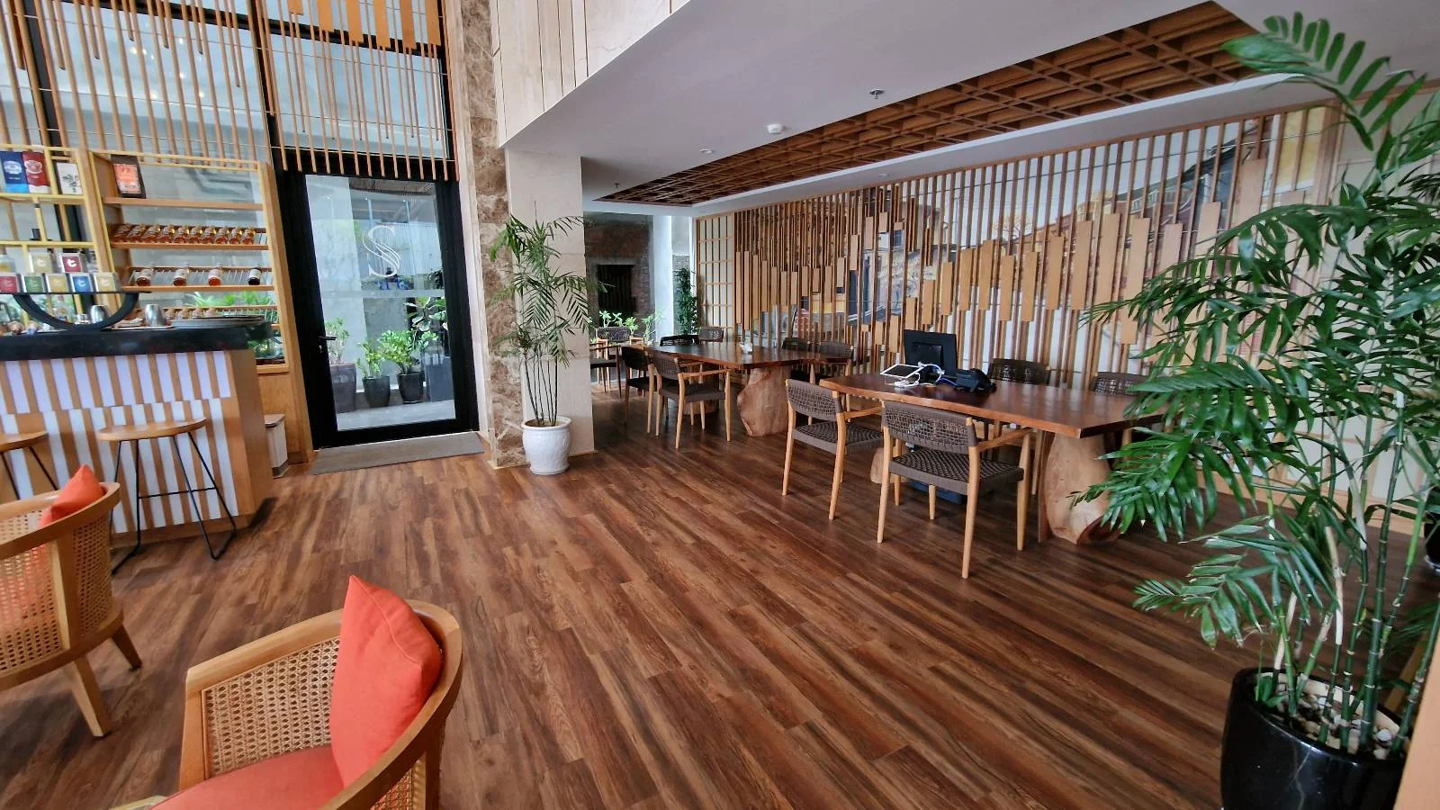 A modern office space with wooden flooring and furniture. There are desks with computers on the right, a potted plant, and a seating area with chairs and a table on the left. Large windows provide natural light.